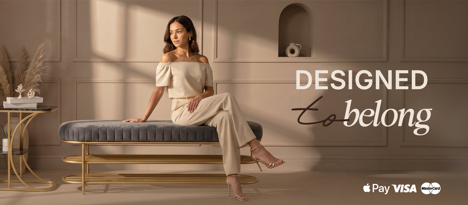 A woman sits on a blue upholstered bench with a gold frame in a warm beige interior. She wears an off-the-shoulder cream top and matching cream pants.