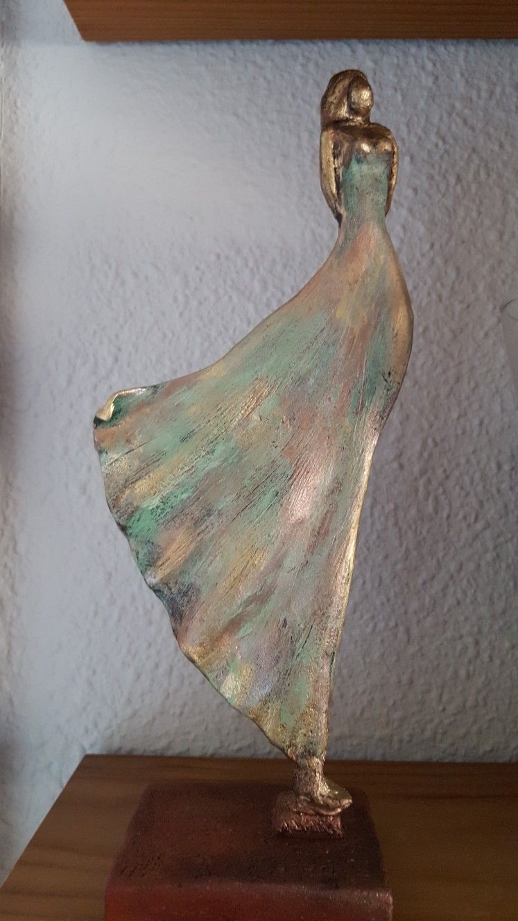 Bronze sculpture of a dancer on a wooden base against a textured white wall.