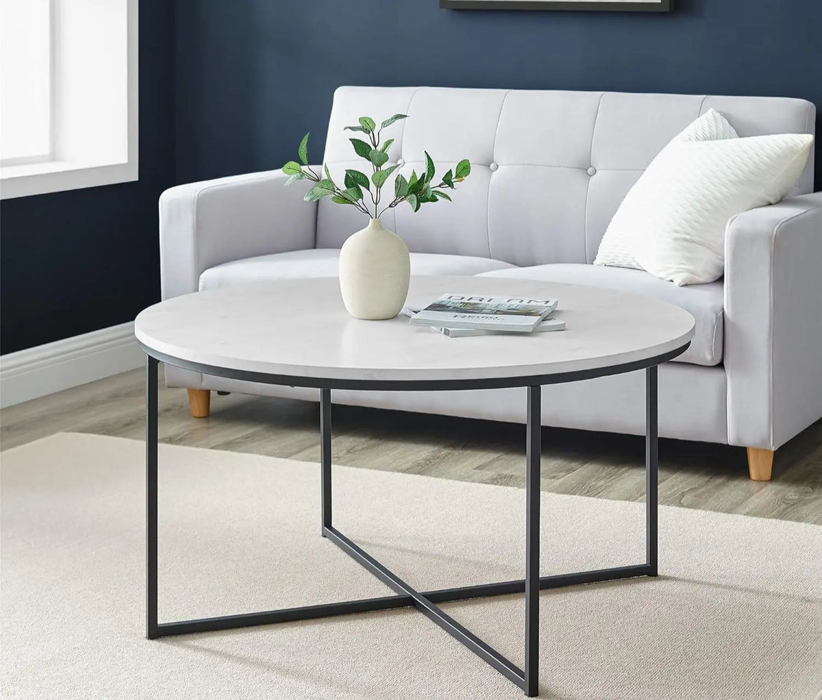 Round coffee table with a vase and books in front of a gray sofa against a blue wall.