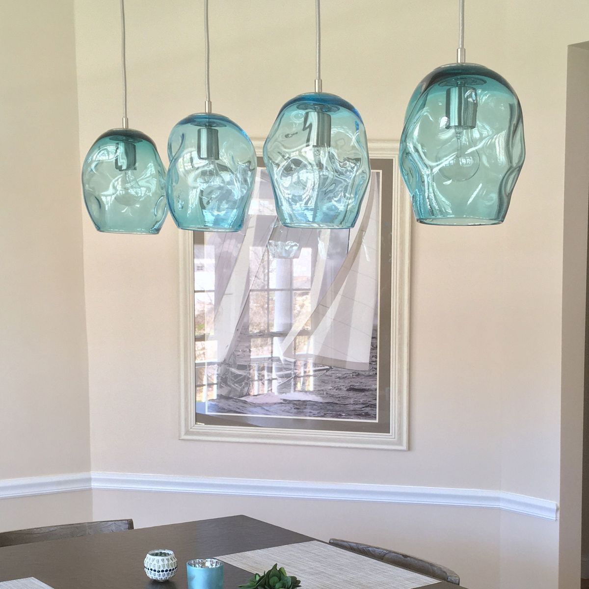Four turquoise pendant lights hanging above a dining table with a neutral wall and framed picture in the background.