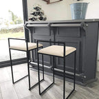 Two bar stools with black metal frames and beige cushioned seats in front of a dark gray bar.