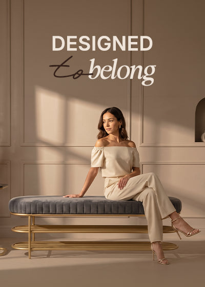 A woman sits on a gray tufted bench with a gold frame in a warm beige room. She wears an off-the-shoulder cream top and matching cream trousers.