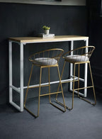 Two gold bar stools with white cushions in front of a wooden table against a dark wall.